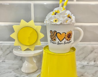 May include: A decorative arrangement featuring a yellow sun-shaped object on a white pedestal, a white mug with the text "Peace Love Sunshine", and a yellow bucket. The mug is topped with white whipped cream, sunflowers, and yellow striped straws.