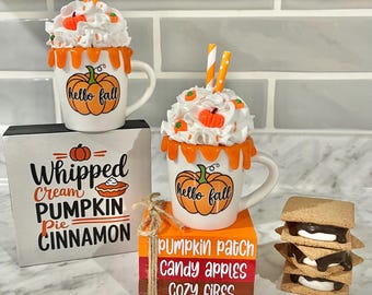 May include: A fall-themed decorative display featuring two mugs with whipped cream and pumpkin accents, and a stack of s'mores. The mugs have "Hello Fall" and pumpkin designs. A wooden block reads "Whipped Cream Pumpkin Pie Cinnamon".