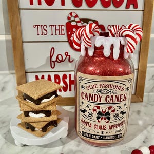 May include: A festive holiday scene with a jar of red glitter, candy cane accents, and a sign that reads "Hot Cocoa." A stack of s'mores sits on a white stand, and a garland of red and white beads adds to the seasonal decor.