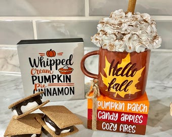 May include: A fall-themed display featuring a mug with whipped cream and a cinnamon stick, a sign that says "Whipped Cream Pumpkin Pie Cinnamon", and three s'mores. The mug says "Hello Fall". There are also stacked blocks that say "Pumpkin Patch Candy Apples Cozy Fires".