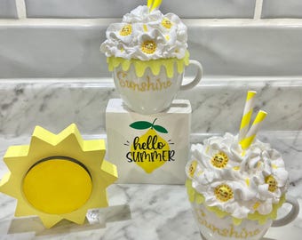 May include: Two white ceramic mugs with yellow and white frosting and yellow smiley faces. The mugs have the word "Sunshine" written on them. The mugs are sitting on a white surface with a yellow wooden sun and a white block with the words "hello summer" and a yellow lemon.