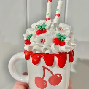May include: A white ceramic mug with a cherry design, topped with whipped cream, cherries, and two cherry-printed straws. The rim of the mug has a red, drippy glaze. A whimsical, decorative item.