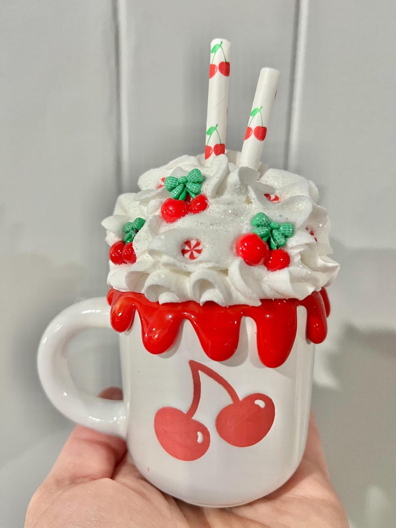 May include: A white ceramic mug with a cherry design, topped with whipped cream, cherries, and two cherry-printed straws. The rim of the mug has a red, drippy glaze. A whimsical, decorative item.