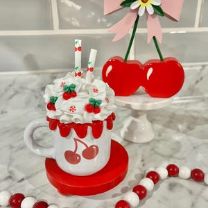 May include: A whimsical cherry-themed display featuring a white mug with cherry graphics, topped with whipped cream and cherry accents. A cherry-topped bow and a red and white beaded garland complete the playful arrangement.