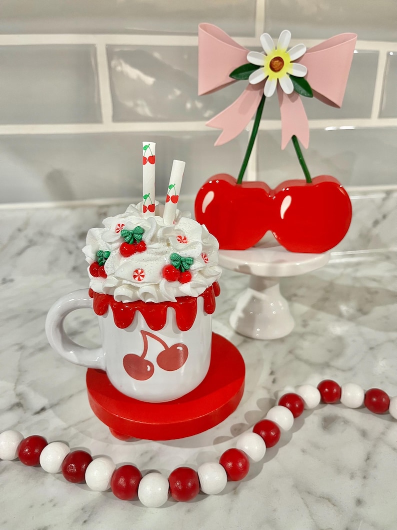 May include: A whimsical cherry-themed display featuring a white mug with cherry graphics, topped with whipped cream and cherry accents. A cherry-topped bow and a red and white beaded garland complete the playful arrangement.