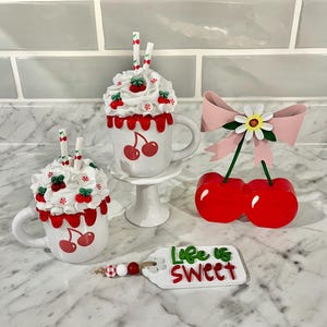May include: A collection of cherry-themed decorative items. Includes two white mugs with cherry designs and whipped cream toppings, a cherry ornament with a pink bow, and a tag that reads "Life is Sweet".