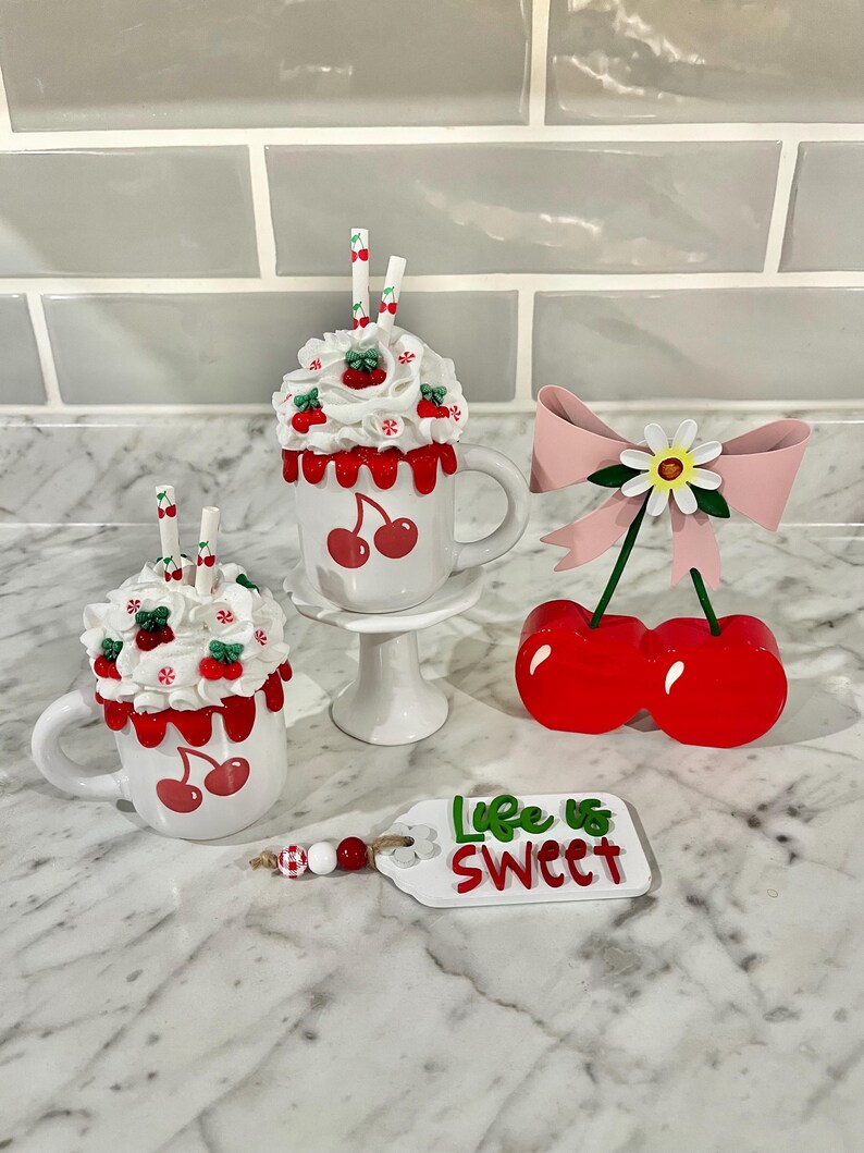 May include: A collection of cherry-themed decorative items. Includes two white mugs with cherry designs and whipped cream toppings, a cherry ornament with a pink bow, and a tag that reads "Life is Sweet".