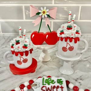May include: A decorative display featuring white mugs with cherry designs, topped with faux whipped cream and cherries. A red cherry ornament and a beaded garland with a tag that reads "Life is Sweet" complete the scene.