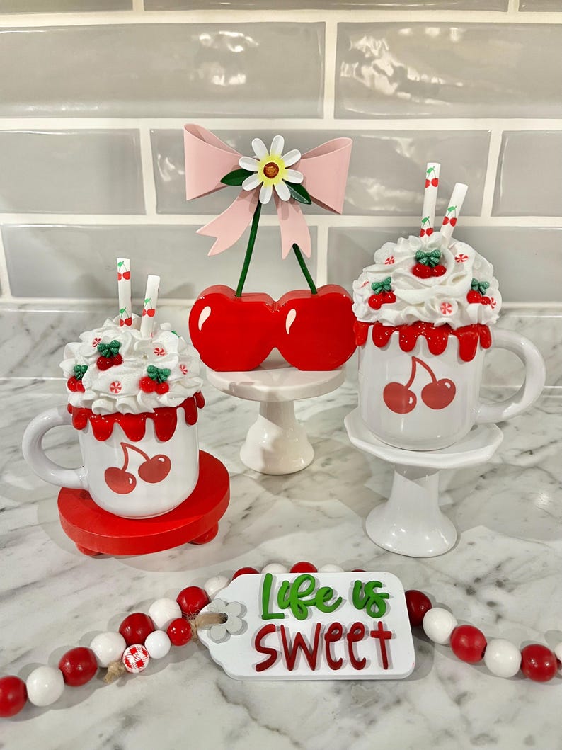 May include: A decorative display featuring white mugs with cherry designs, topped with faux whipped cream and cherries. A red cherry ornament and a beaded garland with a tag that reads "Life is Sweet" complete the scene.