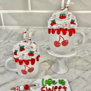 May include: Two white ceramic mugs with cherry designs, topped with whipped cream, cherries, and straws. One mug sits on a small white pedestal. A tag with the words "Life is Sweet" and a beaded garland are also present.