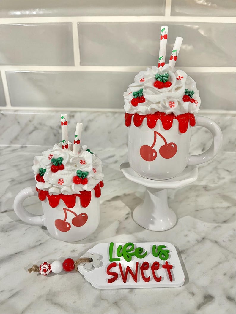 May include: Two white ceramic mugs with cherry designs, topped with whipped cream, cherries, and straws. One mug sits on a small white pedestal. A tag with the words "Life is Sweet" and a beaded garland are also present.