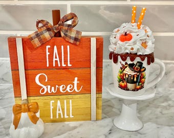 May include: A decorative fall-themed arrangement featuring a wooden sign with the words "FALL Sweet FALL" in white, a ceramic pumpkin, and a mug of whipped cream. The mug is on a white pedestal.