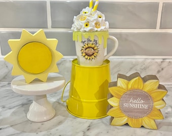 May include: A yellow ceramic mug with a sunflower design and the text "You Are My Sunshine" is sitting on a yellow bucket. A yellow wooden sunflower with the text "hello sunshine" is on the right. A white cake stand with a yellow ceramic sun is on the left.