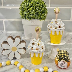 May include: A decorative arrangement featuring a white daisy, a string of yellow and white beads, and two cupcake-shaped containers with white frosting and bee accents. A small jar with a plaid lid and the text "Save Our Bees" is also present.