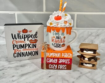 May include: A fall-themed decorative display featuring a mug with whipped cream and a pumpkin, stacked books, a sign, and s'mores. The mug reads "I'm a fall kinda girl." The sign says "Whipped Cream Pumpkin Pie Cinnamon."