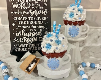 May include: A winter-themed display featuring two decorative mugs with whipped cream and snowflake accents, a sign with text, and a beaded garland. The sign reads "It's a Marshmallow World". A s'more is also present.