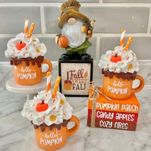 May include: Three ceramic mugs with whipped cream and chocolate drizzle, decorated with orange and white sprinkles. Each mug has a brown handle and says "hello pumpkin". The mugs are arranged on a white surface with a fall-themed gnome and a wooden sign that says "Fall Sweet Fall".