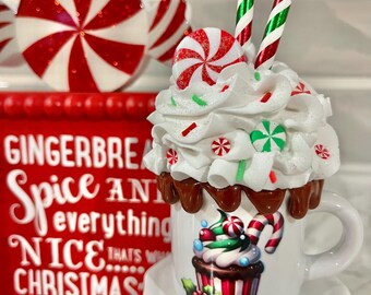 May include: A festive Christmas-themed display featuring a white mug with a cupcake design, topped with whipped cream, candies, and candy canes. A red sign with the text "Gingerbread Spice AND everything NICE...THAT'S WHAT CHRISTMAS" is also visible.