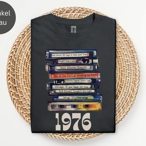 Könnte beinhalten: Dunkelgraues T-Shirt mit einem Stapel Kassetten, auf denen Songtitel wie "Bohemian Rhapsody" und "Disco Lady" stehen. Die Zahl "1976" ist darunter in Weiß aufgedruckt. Das Shirt wird auf einer gewebten Matte präsentiert.