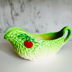 May include: A ceramic gravy boat with a lettuce leaf design. The boat is primarily light green with a textured surface, a red tomato accent, and a curved handle. The interior is a lighter shade of green.