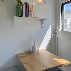 May include: A wall-mounted wooden shelf holding a row of books, positioned above a square wooden table. A white ceramic vase sits on the table surface. A glass pendant light hangs from the ceiling, and a window is visible on the right side.