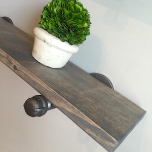 May include: A dark gray stained wooden shelf mounted with black industrial metal pipe brackets. A small, round, vibrant green topiary plant sits in a weathered white ceramic pot on the surface of the wooden shelf, which is secured to the wall.