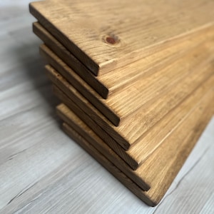 May include: A stack of unfinished wooden planks. The planks are a light brown color with visible wood grain and knots. The edges are cleanly cut, and the planks are stacked neatly, suggesting they are ready for a woodworking project or home decor.
