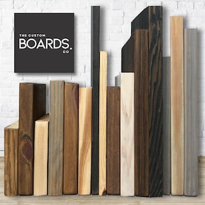 Op de afbeelding: Een verzameling houten planken in verschillende tinten bruin, grijs en wit. De planken zijn verticaal gerangschikt en hebben allemaal verschillende breedtes. De planken zijn gemaakt van gerecycled hout en hebben een rustieke uitstraling. De tekst "THE CUSTOM BOARDS CO." is zichtbaar in de linkerbovenhoek van de afbeelding.
