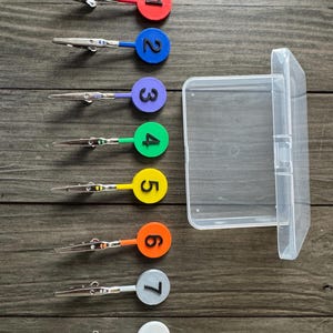 May include: A set of eight numbered alligator clips with a clear plastic storage case. Each clip has a different coloured circular top with a number from one to eight. The clips are arranged on a wooden surface.