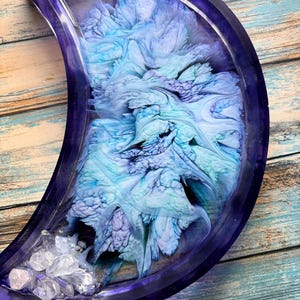 May include: A crescent moon-shaped tray made of dark purple resin. The center features swirling patterns of light blue, white, and soft purple ink. A cluster of clear, jagged, crystal-like stones is embedded at the bottom tip of the tray.