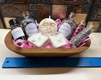 May include: A wooden bowl filled with various spiritual items. It contains small glass bottles with corks, candles, and a moon-shaped candle. The bottles are labeled with terms like "Prosperity" and "Divination". Pink paper and a blue ruler are also in the bowl.