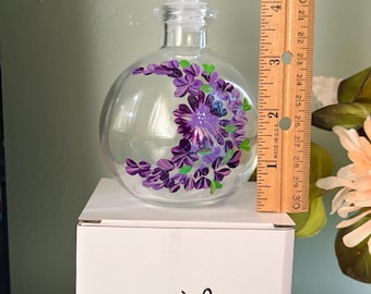 May include: A clear glass bottle with a spherical body and a round stopper, featuring hand-painted purple flowers in a crescent moon shape. A wooden ruler placed next to the bottle indicates a height of approximately 5.5 inches.