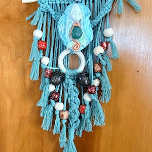 May include: A turquoise macrame wall hanging featuring a central ceramic figure with blue hair and a green stone. The design includes white, red, and brown beads, along with a white ring and black decorative elements. The piece is suspended from a wooden dowel.
