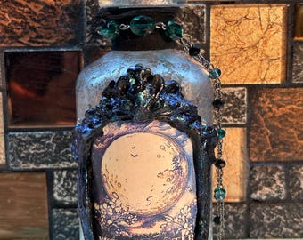 Könnte beinhalten: Eine silberne Glasflasche mit einem schwarzen und grünen verzierten Rahmen um ein Etikett mit der Aufschrift "MOON WATER". Die Flasche hat einen Korkverschluss und eine Perlenkette mit grünen und schwarzen Perlen.