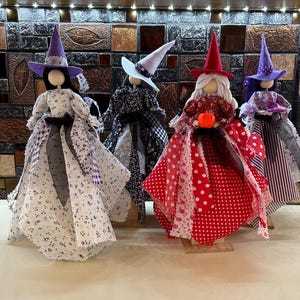 May include: Four handmade witch dolls with fabric dresses and pointed hats. The dolls have different colored hats and dresses, including purple, white, black and white, and red. One doll holds a small orange pumpkin.