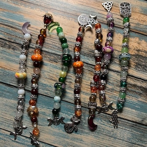 May include: A collection of beaded bookmarks with various charms. The bookmarks feature a variety of colorful beads, including orange, green, red, and purple. Charms include moons, cats, and other mystical symbols. The bookmarks are arranged on a wooden surface.