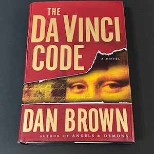 May include: A hardcover book titled "The Da Vinci Code" by Dan Brown. The book has a red cover with gold lettering. The cover features an image of eyes and part of a face. The author is also the author of "Angels & Demons".
