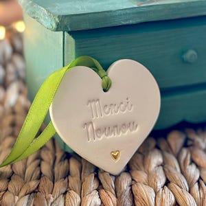 Op de afbeelding: Een wit hartvormig keramisch label met de woorden "Merci Nounou" en een klein gouden hart. Het label is bevestigd aan een groen lint. Op de achtergrond een turquoise geschilderde houten kist met een bloemmotief.