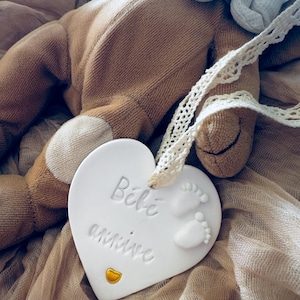 Może przedstawiać: Biała ceramiczna ozdoba w kształcie serca z wytłoczonym napisem "Bébé arrive" i dwoma małymi odciskami stóp dziecka. Na dole znajduje się małe złote serduszko, a u góry przymocowana jest kremowa koronkowa wstążka do zawieszenia ozdoby.