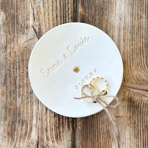 Peut inclure: Un porte-anneaux en céramique blanche avec les noms "Emma & Louis" et la date "31.08.24" en relief. Un petit cœur doré et deux bagues dorées sont également présents. Les bagues sont attachées avec de la ficelle. Le plat est posé sur une surface en bois.