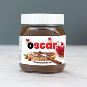 Puede incluir: Un tarro de vidrio de crema de chocolate y avellanas con tapa blanca. La etiqueta es blanca con corazones rojos y el texto "oscar" en letras negras y rojas. La etiqueta también muestra una rebanada de pan con crema de chocolate y un cuchillo. El texto "OPTIONAL MESSAGE HERE" está impreso debajo del pan.