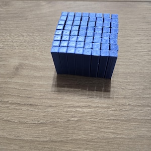 Op de afbeelding: Een vierkant blok bestaande uit vele kleine, verticale, blauwe rechthoekige prisma's gerangschikt in een rasterpatroon. Het object heeft een uniforme matte blauwe afwerking op alle zichtbare boven- en zijkanten, wat een solide, compacte en geometrische structuur vormt.