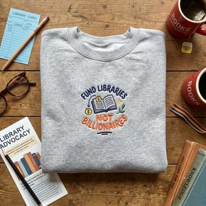 May include: A heather gray sweatshirt with the embroidered text "FUND LIBRARIES NOT BILLIONAIRES" in blue, orange, and yellow. The design includes an open book, dollar signs, and leaves. The image also shows books, mugs, and a library card.