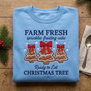 May include: A light blue crewneck sweatshirt featuring embroidered text that reads FARM FRESH sprinkles frosting cake and Ready to Eat CHRISTMAS TREE, with three brown gingerbread trees decorated with red bows and green sprinkles.
