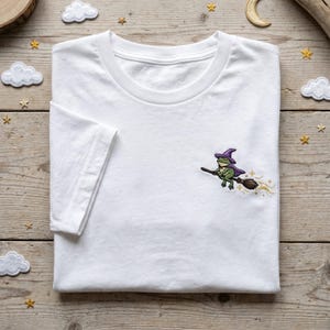 May include: White t-shirt featuring an embroidered design of a green frog in a purple witch hat and cape, riding a broom with gold sparkles. The shirt is folded on a wooden surface.