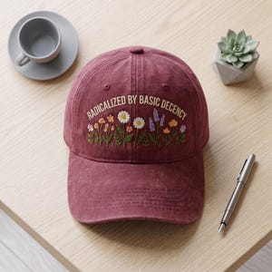 May include: A maroon baseball cap featuring an embroidered design of colourful wildflowers and the text "RADICALIZED BY BASIC DECENCY" in a light tan font. The cap has a structured crown, a curved brim with stitching, and a button on top.