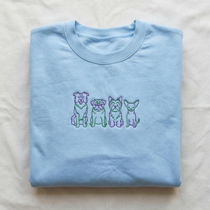 Peut inclure: Un sweat-shirt bleu clair avec un motif brodé de quatre races de chiens différentes en vert et violet. Les chiens sont disposés en rangée sur la poitrine du vêtement plié. Le sweat-shirt est en matière douce.