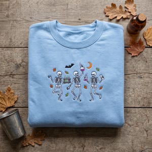 May include: A light blue crewneck sweatshirt featuring an embroidered graphic of three dancing skeletons. The skeletons are holding laboratory glassware, including test tubes and beakers with purple liquid, surrounded by a bat, a crescent moon, and leaves.