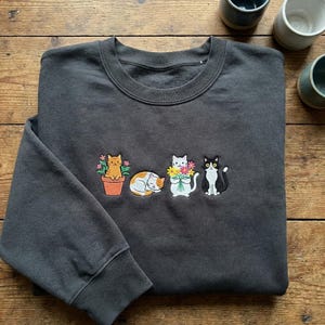 May include: Dark gray sweatshirt with embroidered cats. The design features a ginger cat in a pot, a sleeping orange and white cat, a white cat holding flowers, and a black and white cat. The sweatshirt has a crew neck and long sleeves.
