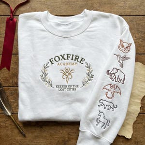 May include: White sweatshirt with embroidered design. The front features the text "FOXFIRE ACADEMY" within a wreath, and "KEEPER OF THE LOST CITIES" below. The right sleeve displays embroidered animals: owl, hummingbird, elephant, dragon, panther, and unicorn.